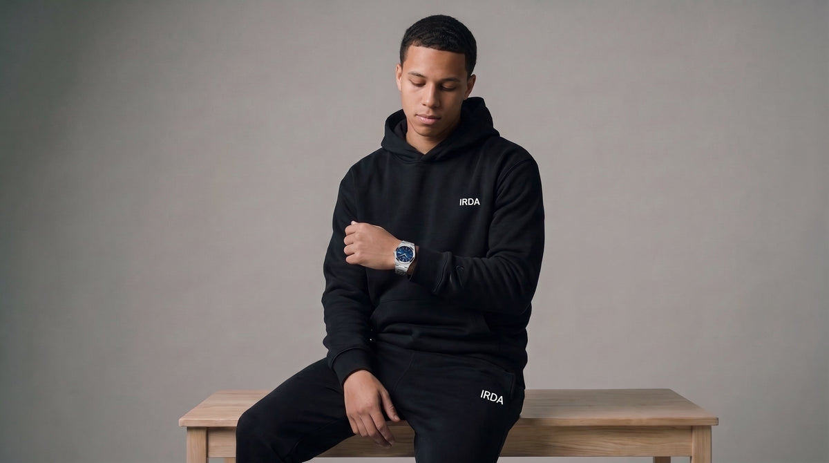 Man seated on bench in black IRDA hoodie and joggers checking a stainless steel watch